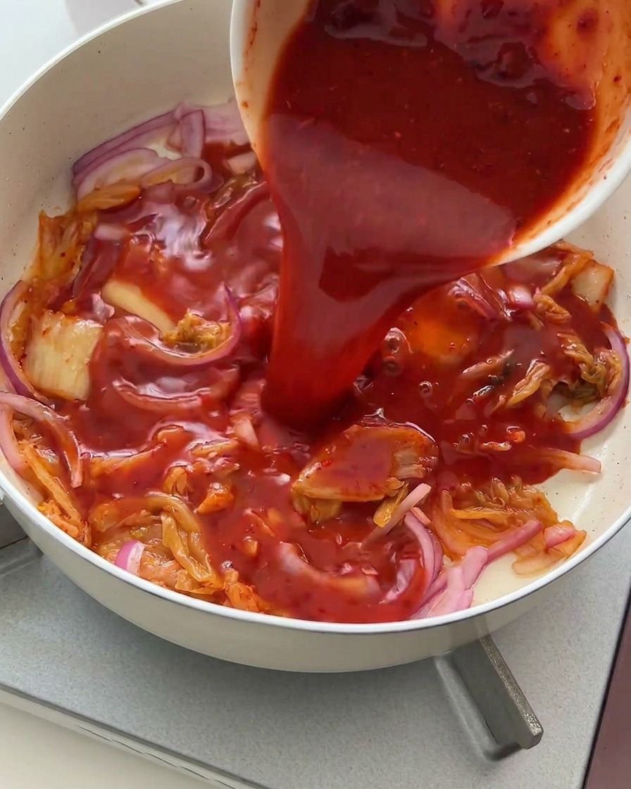 볶은 김치와 양파 위에 그릇에 담긴 붉은 양념장을 붓는 모습.