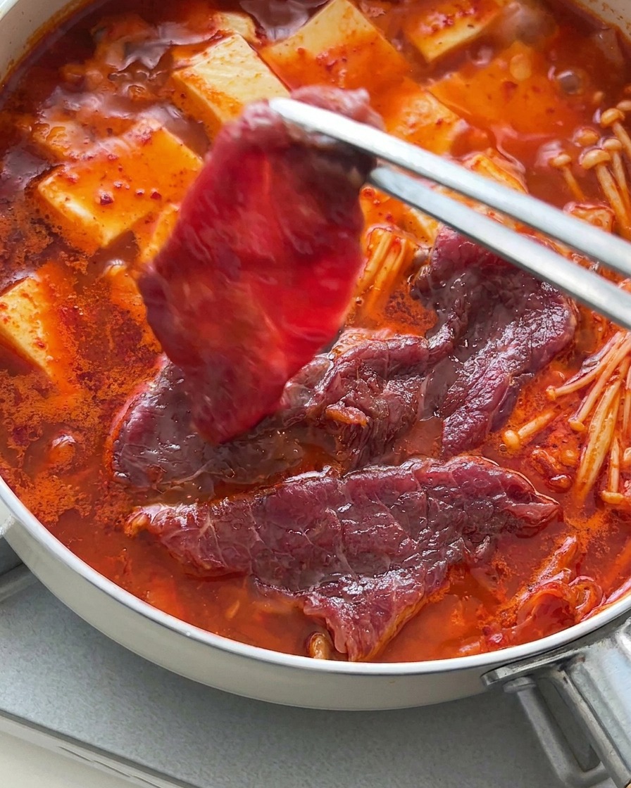 끓는 찌개 위에 집게로 얇은 생소고기 조각들을 펴서 올리는 모습.