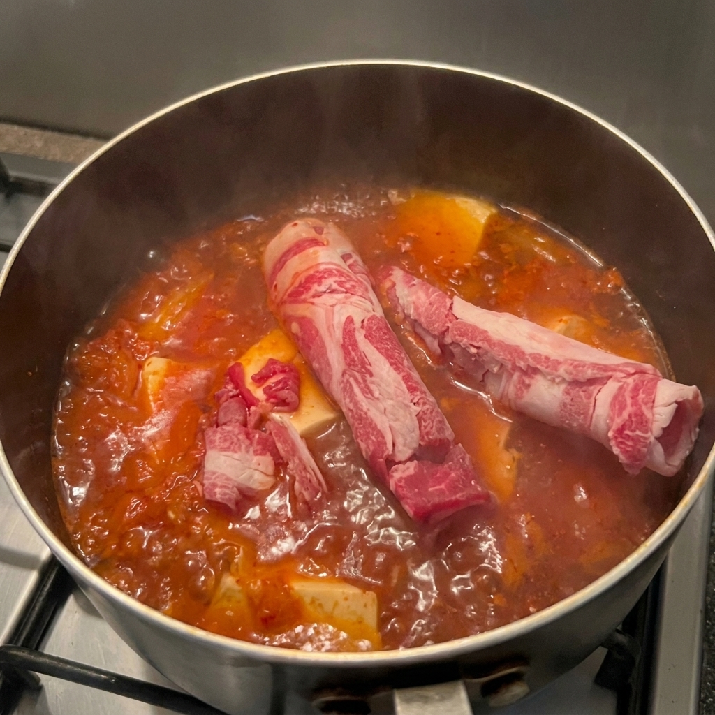 끓는 붉은 김치찌개 위로 마블링이 있는 얇게 썬 생고기를 얹는 모습