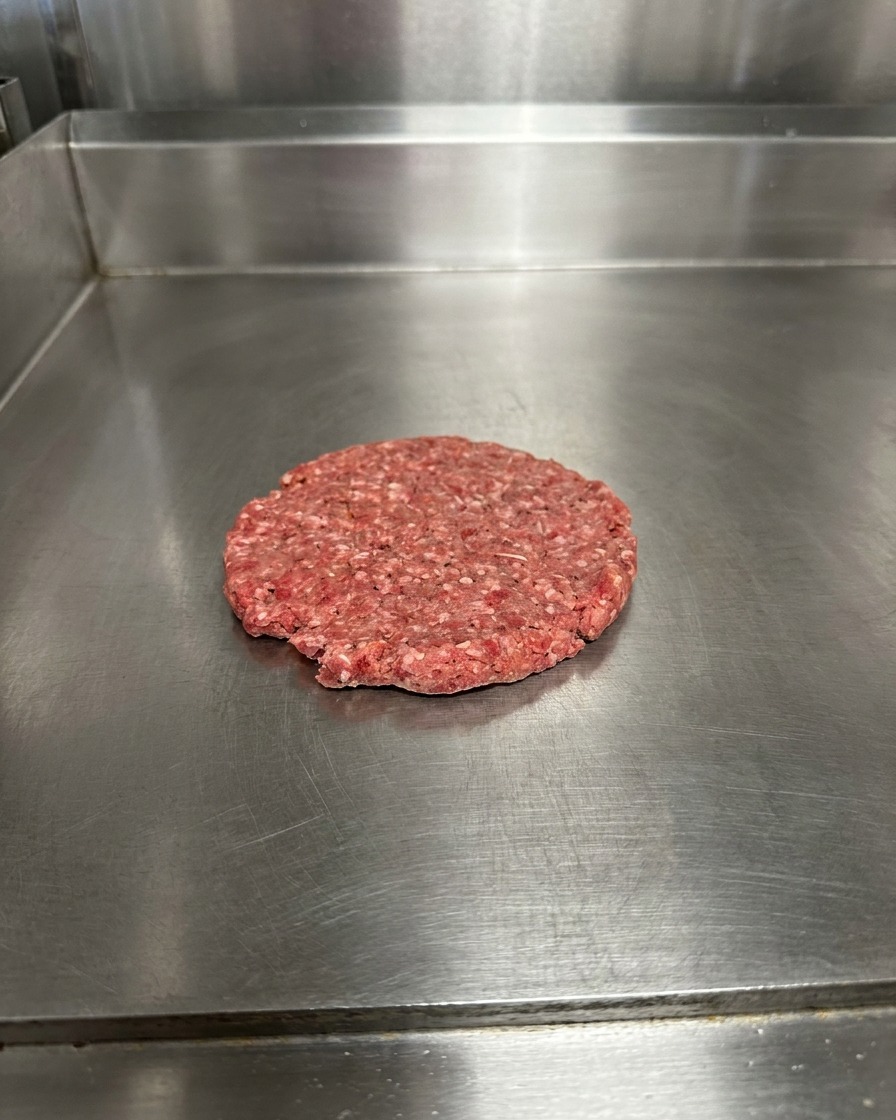 그리들 위에서 납작하게 펴진 소고기 패티에 소금과 후추가 뿌려져 있는 모습.