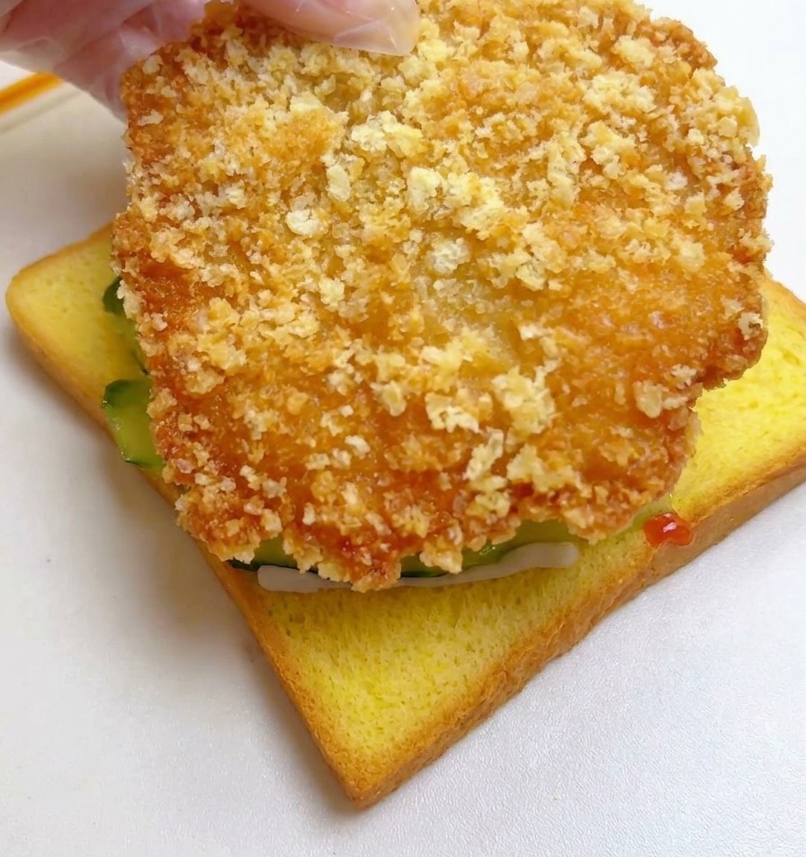 오이가 올라간 식빵 위에 큼직한 황금빛 치킨 필레를 올리는 모습