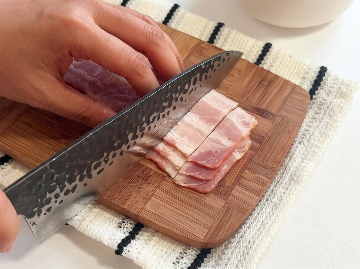 도마 위에서 큰 칼을 사용하여 베이컨 줄을 작은 조각으로 써는 모습.