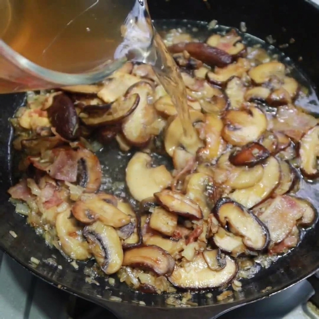 베이컨과 버섯이 있는 팬에 치킨 스톡을 붓는 모습.