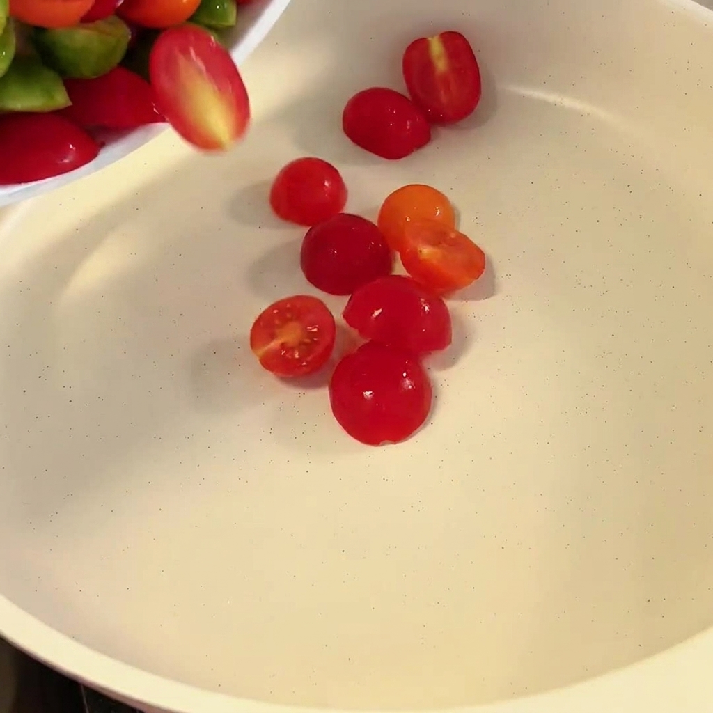 반으로 자른 빨간색과 노란색 방울토마토가 흰색 프라이팬으로 들어가는 모습.