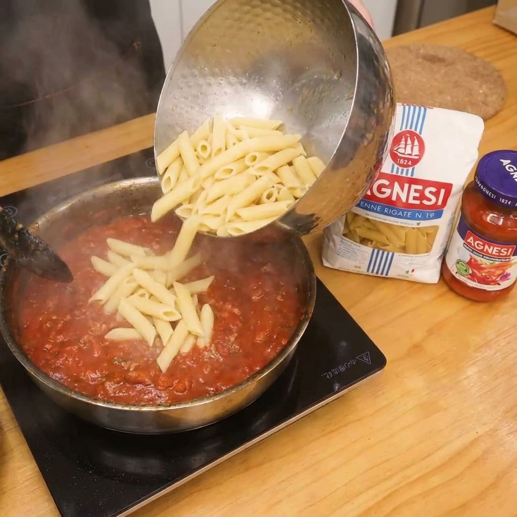 토마토 베이컨 소스가 담긴 팬에 삶은 펜네 파스타를 넣고 섞는 모습.