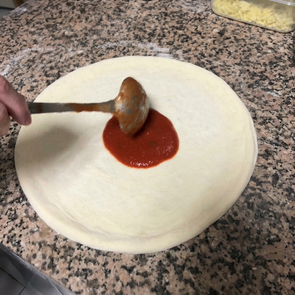 금속 국자를 이용해 원을 그리며 도우에 붉은 토마토 소스를 바르는 모습.