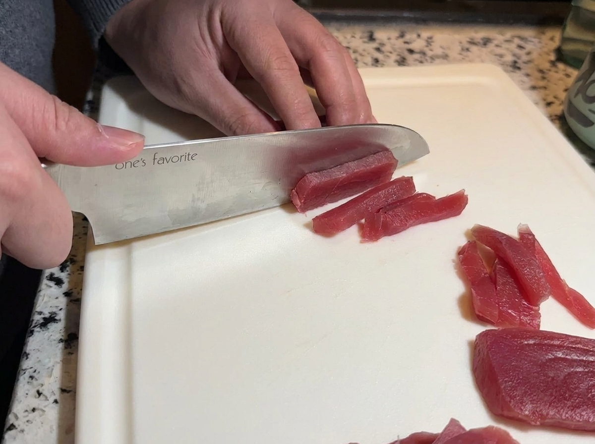 하얀 도마 위에서 날카로운 식칼로 붉은 생참치를 길게 써는 모습.
