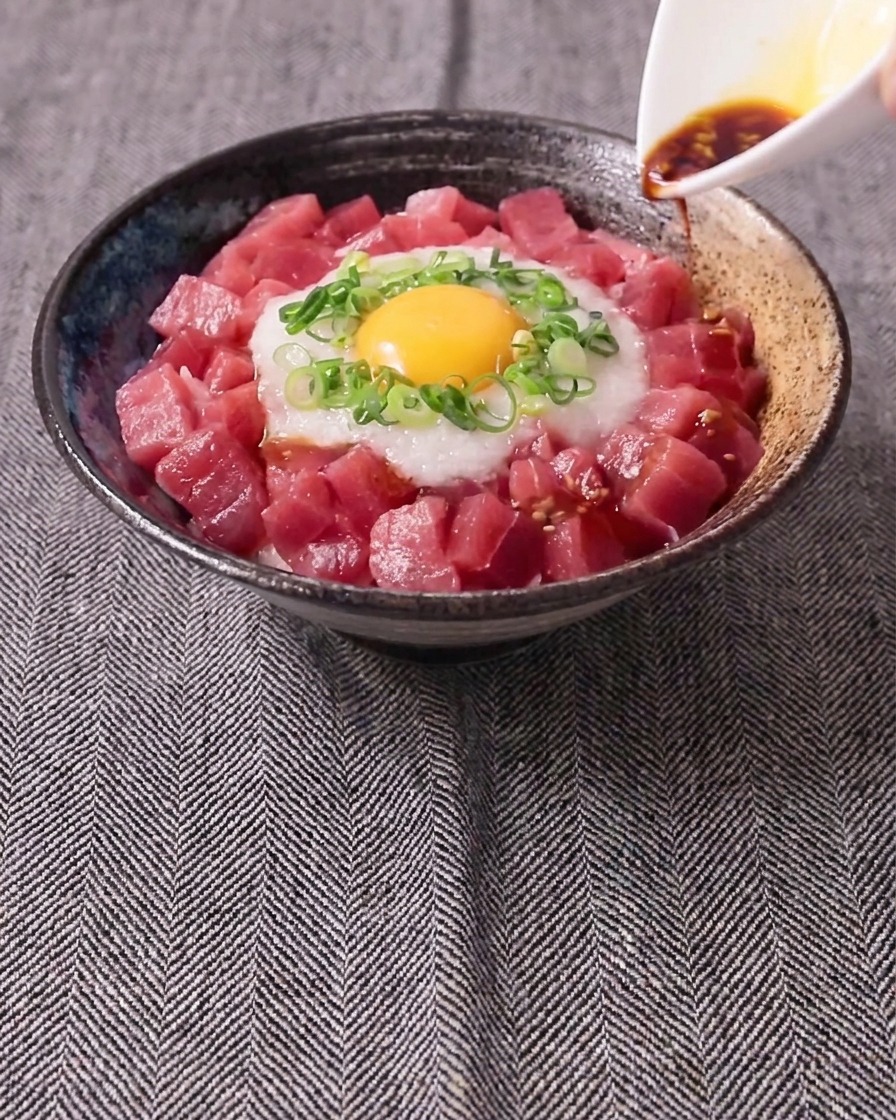 작은 흰색 저그에서 간장 소스를 생참치, 간 마, 쪽파, 생 노른자가 담긴 그릇 위에 붓는 모습.