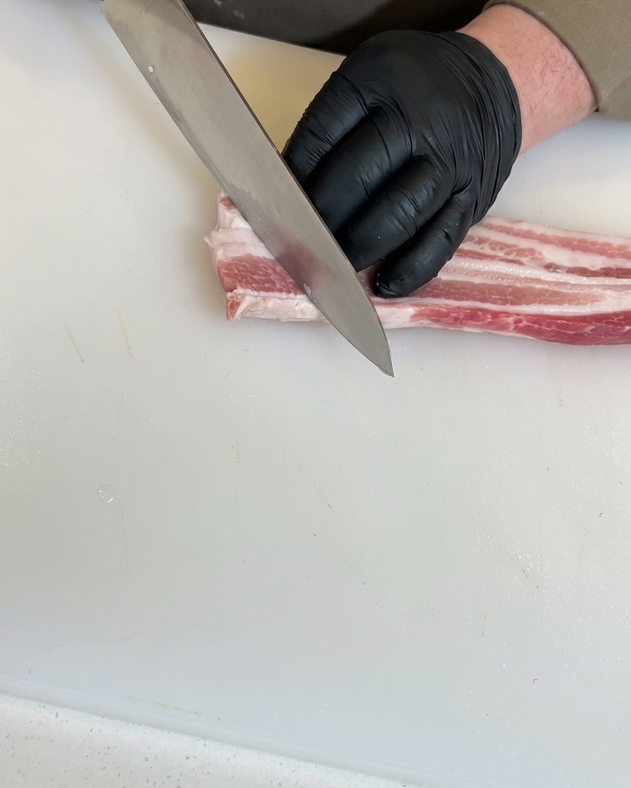 검은 장갑을 낀 손이 흰 도마 위에서 생삼겹살에 대각선으로 깊은 칼집을 넣고 있는 모습.