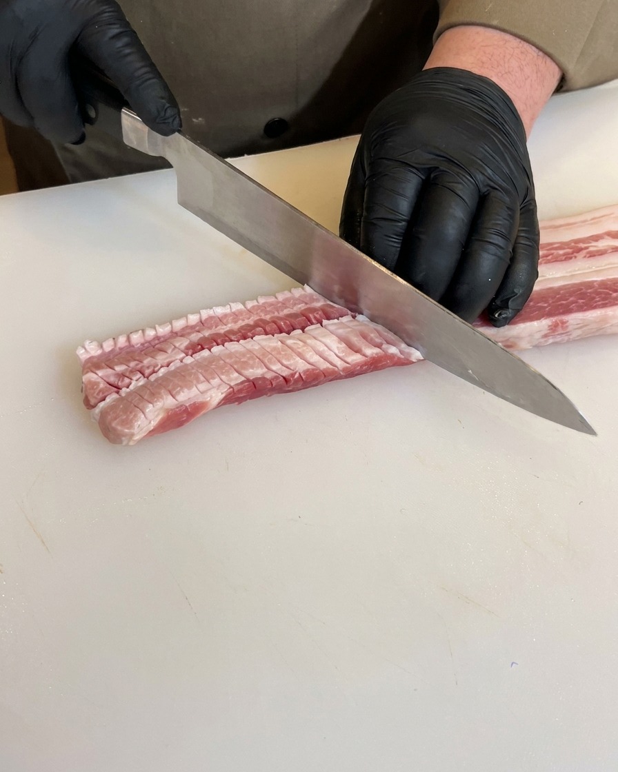 검은 장갑을 낀 손이 삼겹살을 뒤집어 반대편에도 격자 모양으로 깊은 칼집을 내는 모습.