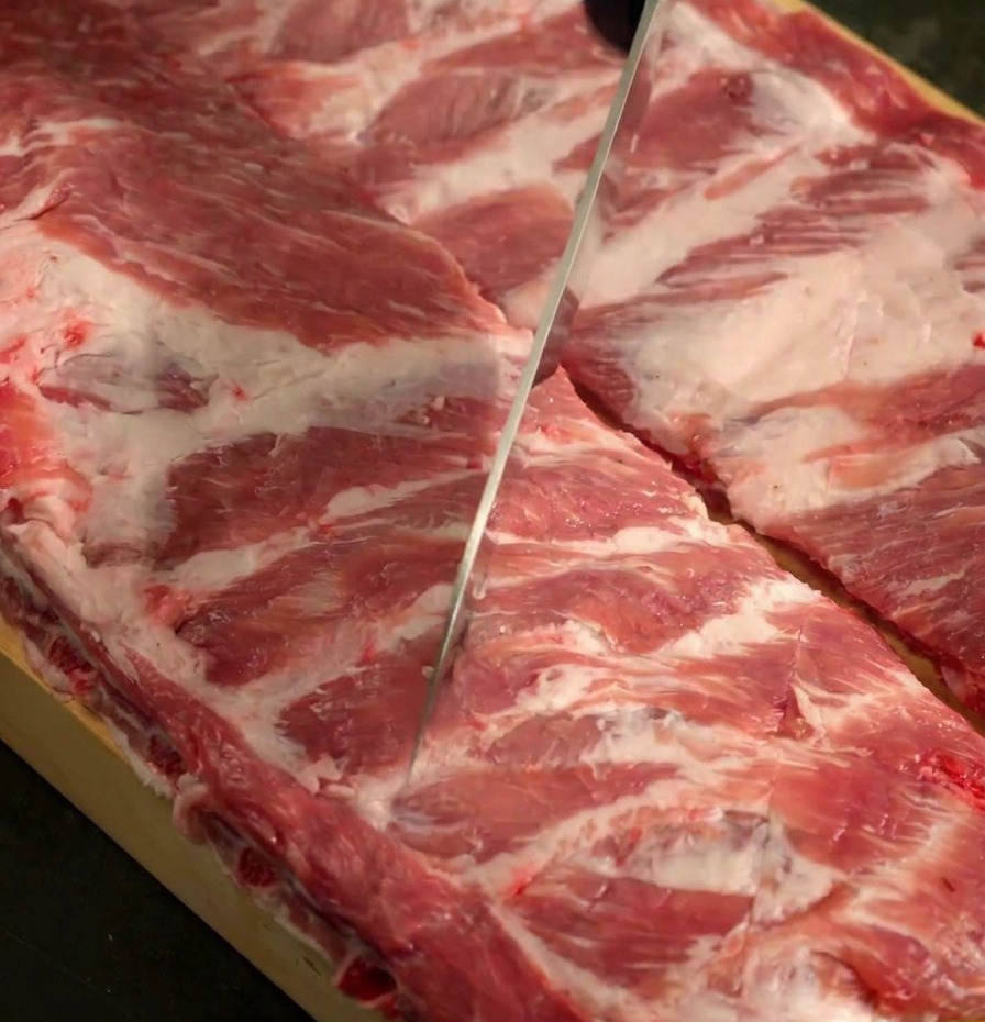 나무 도마 위에서 날카로운 칼로 돼지 등갈비 두 대의 표면에 칼집을 내는 모습.
