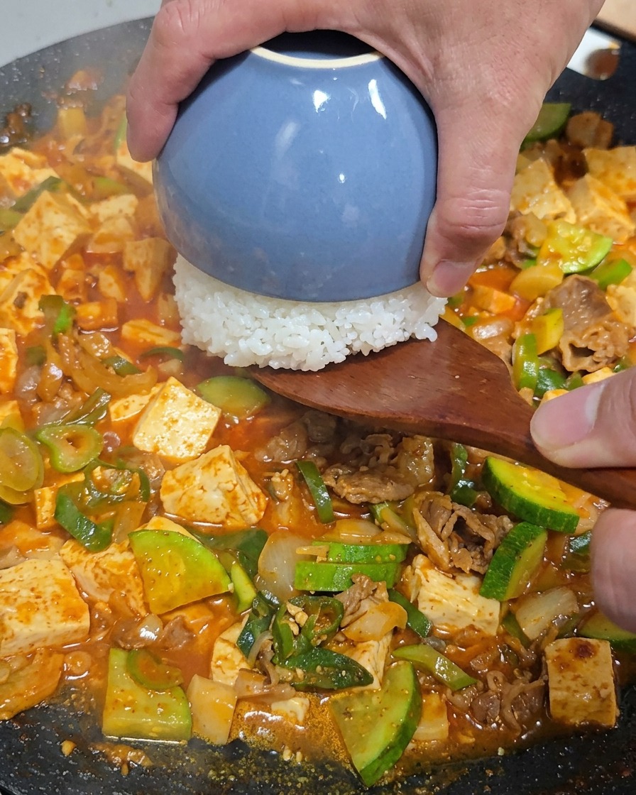 주걱을 사용하여 밥그릇에 담긴 흰 쌀밥을 찌개 중앙에 뒤집어 넣는 모습.