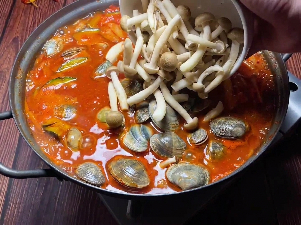 끓고 있는 빨간 김치 찌개에 바지락과 버섯들을 넣는 모습.