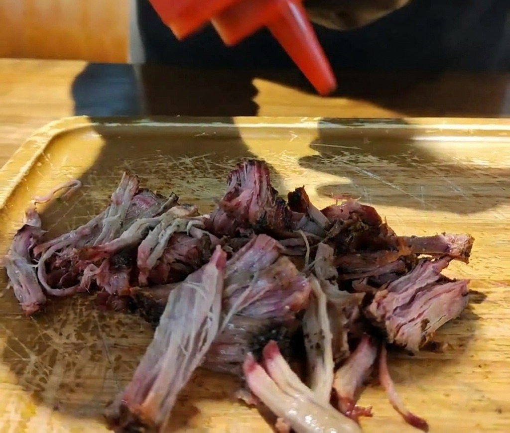 나무 도마 위 잘게 찢은 훈제 돼지고기에 빨간 병에서 진한 바비큐 소스를 뿌리는 모습.