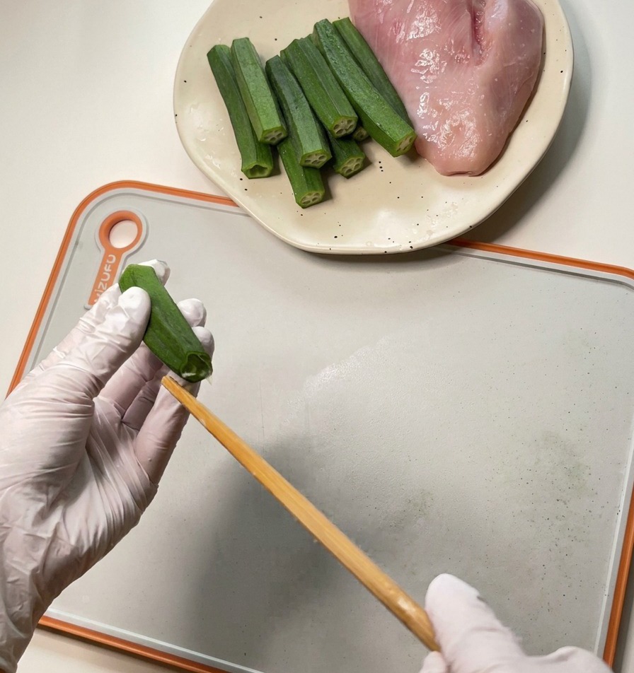 흰 장갑을 낀 사람이 나무 젓가락을 사용하여 오크라 내부의 씨앗과 과육을 밀어내는 모습.