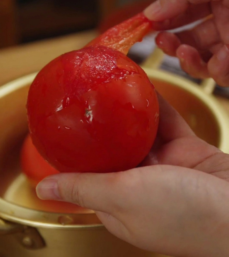 부드러워진 토마토에서 얇고 투명한 껍질을 손으로 벗겨내고 있는 클로즈업 사진.