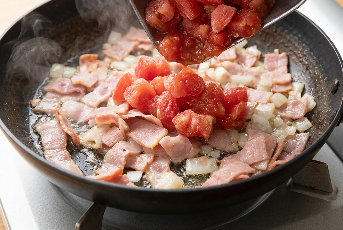 팬에서 베이컨과 양파가 익어가는 중에 다진 토마토를 추가하는 장면.
