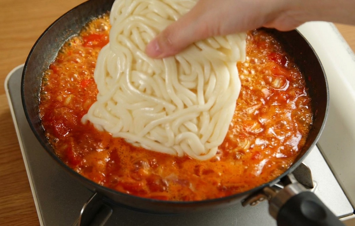 끓고 있는 빨간 토마토 소스 팬에 하얀 우동 사리를 넣는 모습.