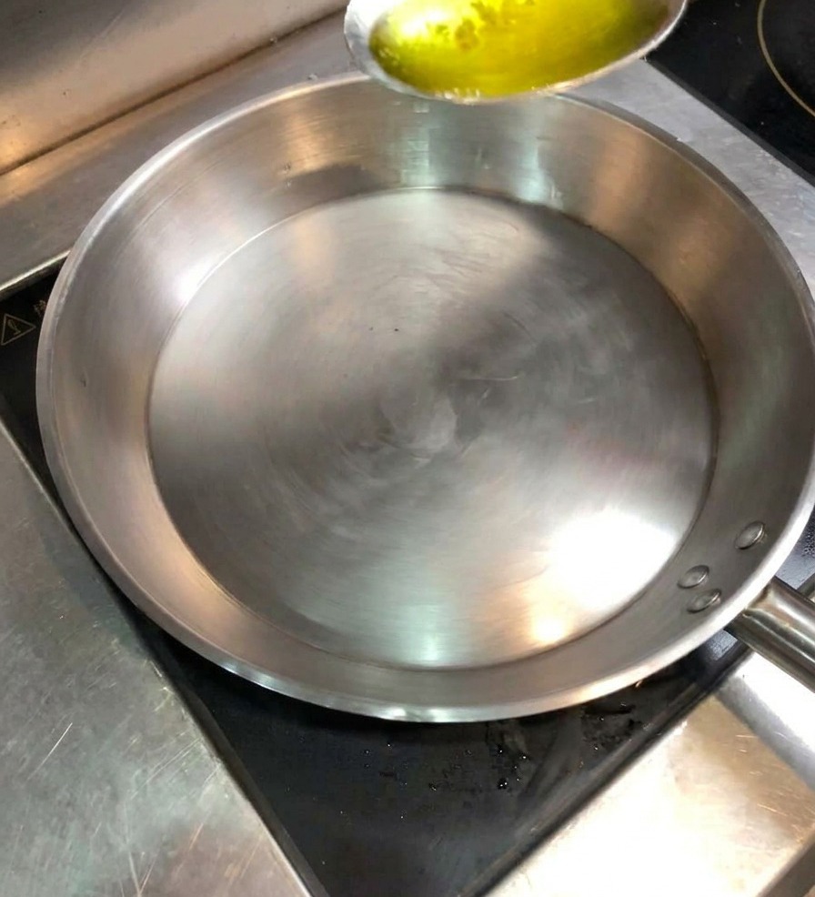 가열된 스테인리스 팬에 금빛 식용유를 숟가락으로 붓고 있는 모습.