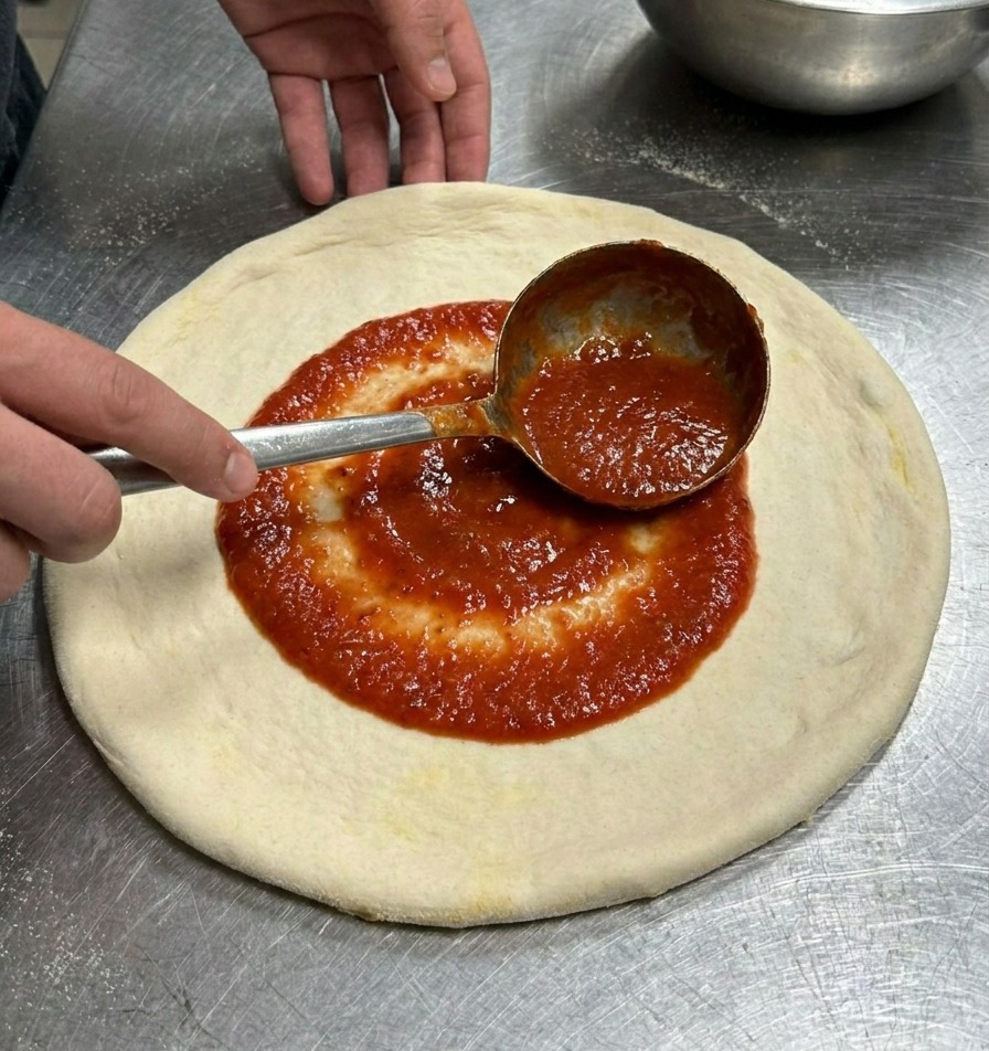 국자를 이용해 빨간 토마토 소스를 원형 도우 중앙부터 나선형으로 펴 바르는 모습.