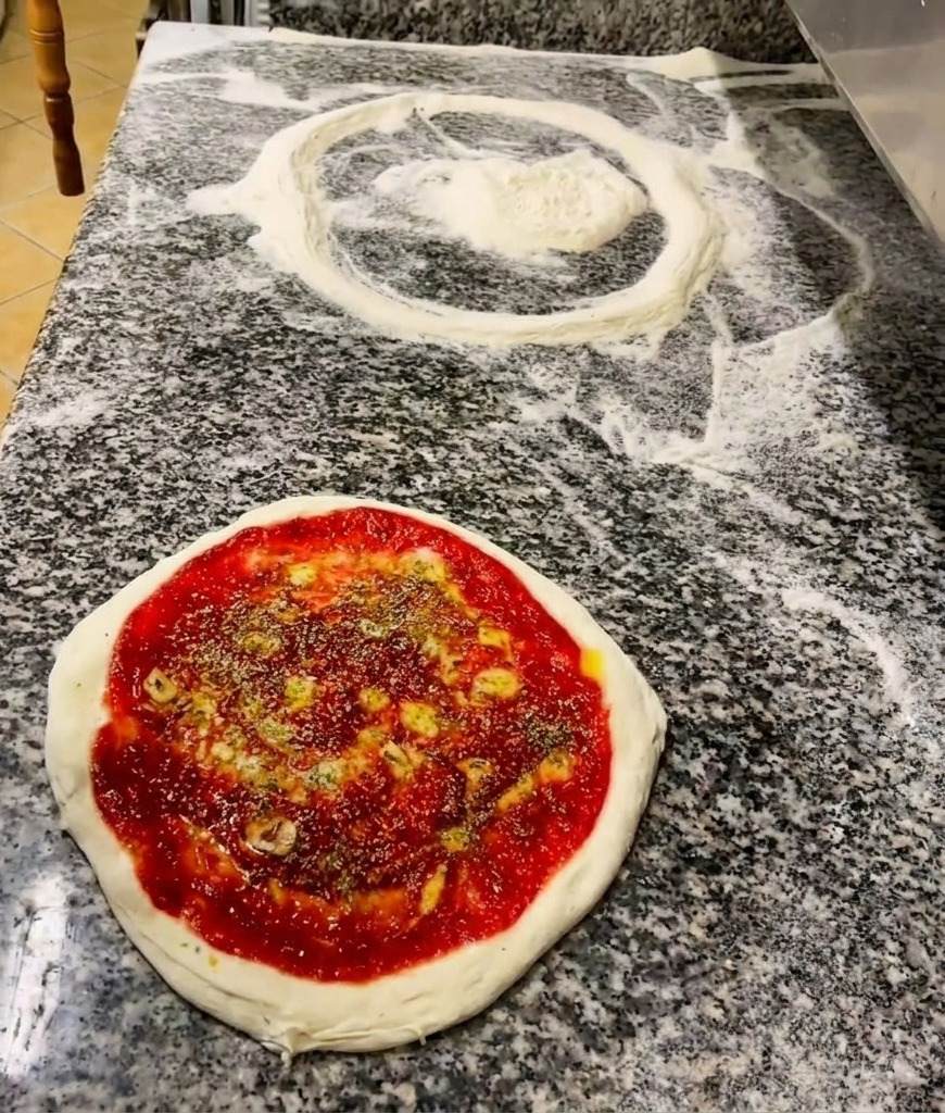 토마토 소스와 올리브유가 뿌려진 피자 도우 위에 초록색 드라이 허브를 뿌리는 모습