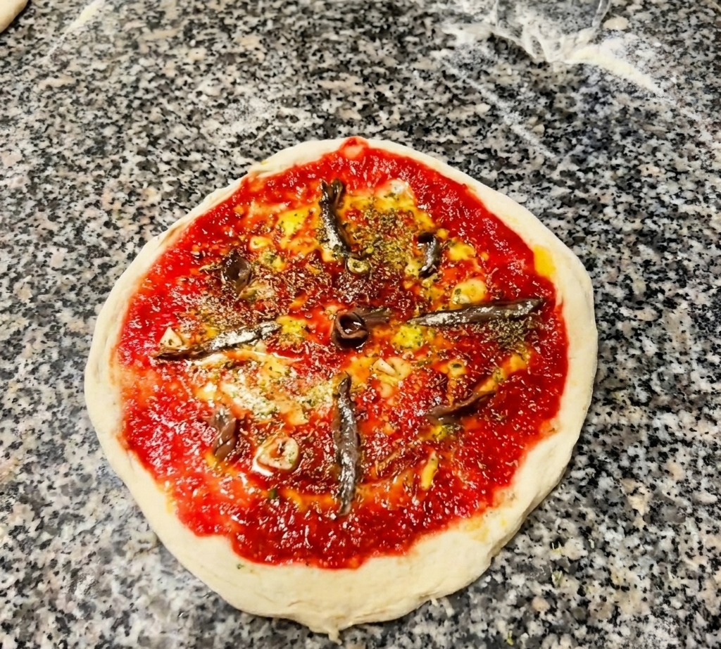 토마토 소스와 허브가 뿌려진 도우 위에 은빛 앤초비 필렛을 바퀴살 모양으로 배치한 모습