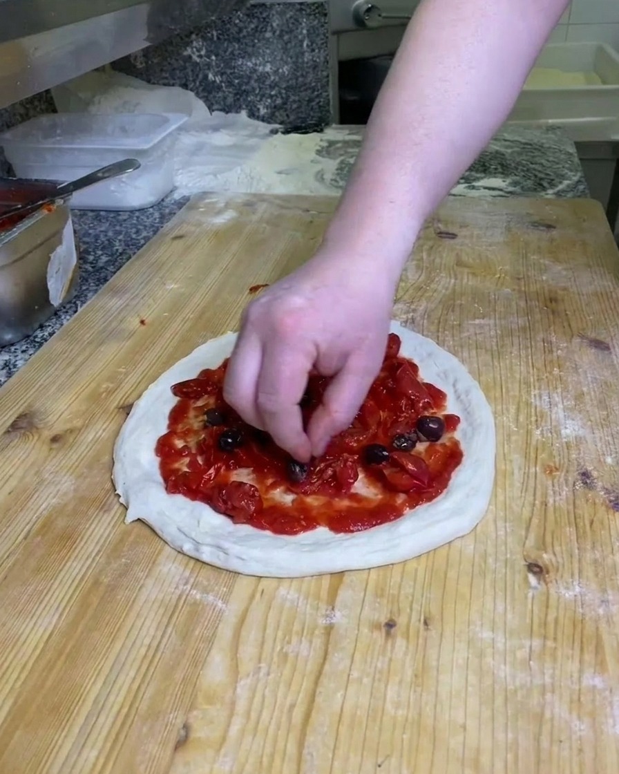 밀가루가 뿌려진 작업대 위 토마토가 덮인 피자 도우 위에 통 블랙 올리브를 배치하는 모습
