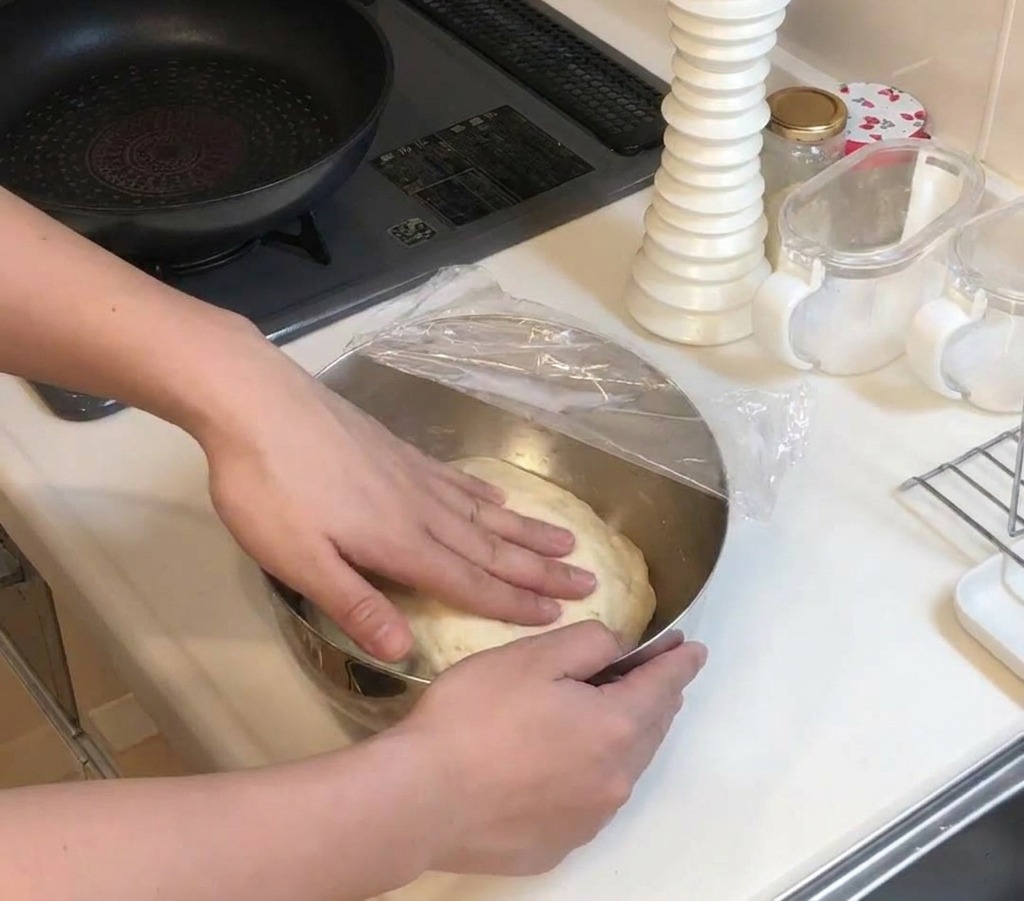 비닐 랩으로 덮인 금속 볼 안에서 부풀어 오른 둥근 반죽을 손으로 눌러보는 모습.