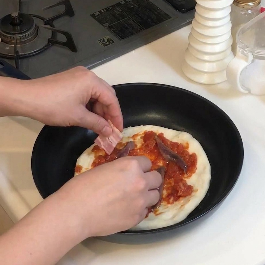 토마토소스와 엔초비가 올려진 피자 위에 생 베이컨 조각을 올리는 모습.