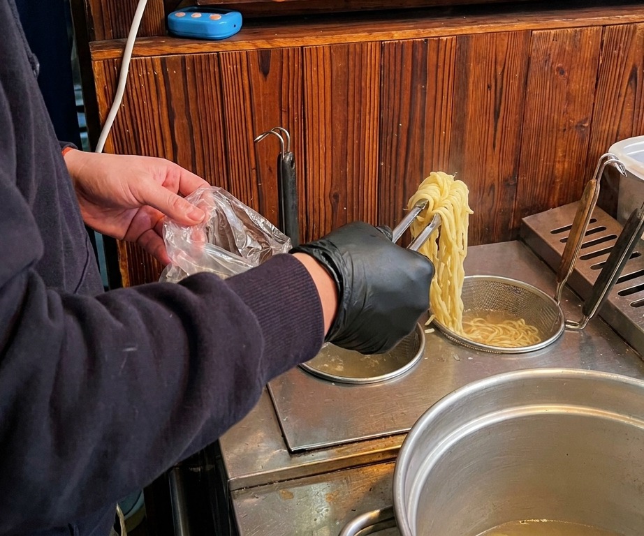 검은 장갑을 낀 조리사가 집게로 끓는 물에서 노란 라멘 면을 건져 올려 그릇에 담으려는 모습.
