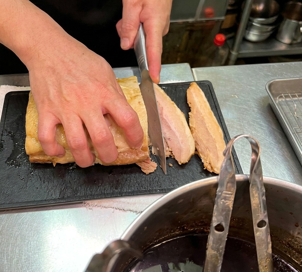 검은색 도마 위에서 부드럽게 조려진 돼지고기 차슈를 두툼하게 썰고 있는 셰프의 모습.