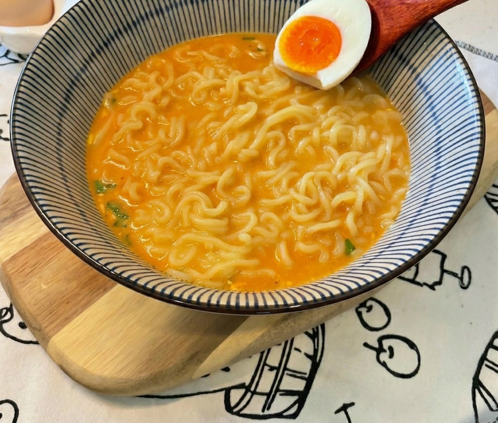 선명한 주황색 노른자가 보이는 반숙 계란을 나무 숟가락으로 라면 위에 올리는 모습.