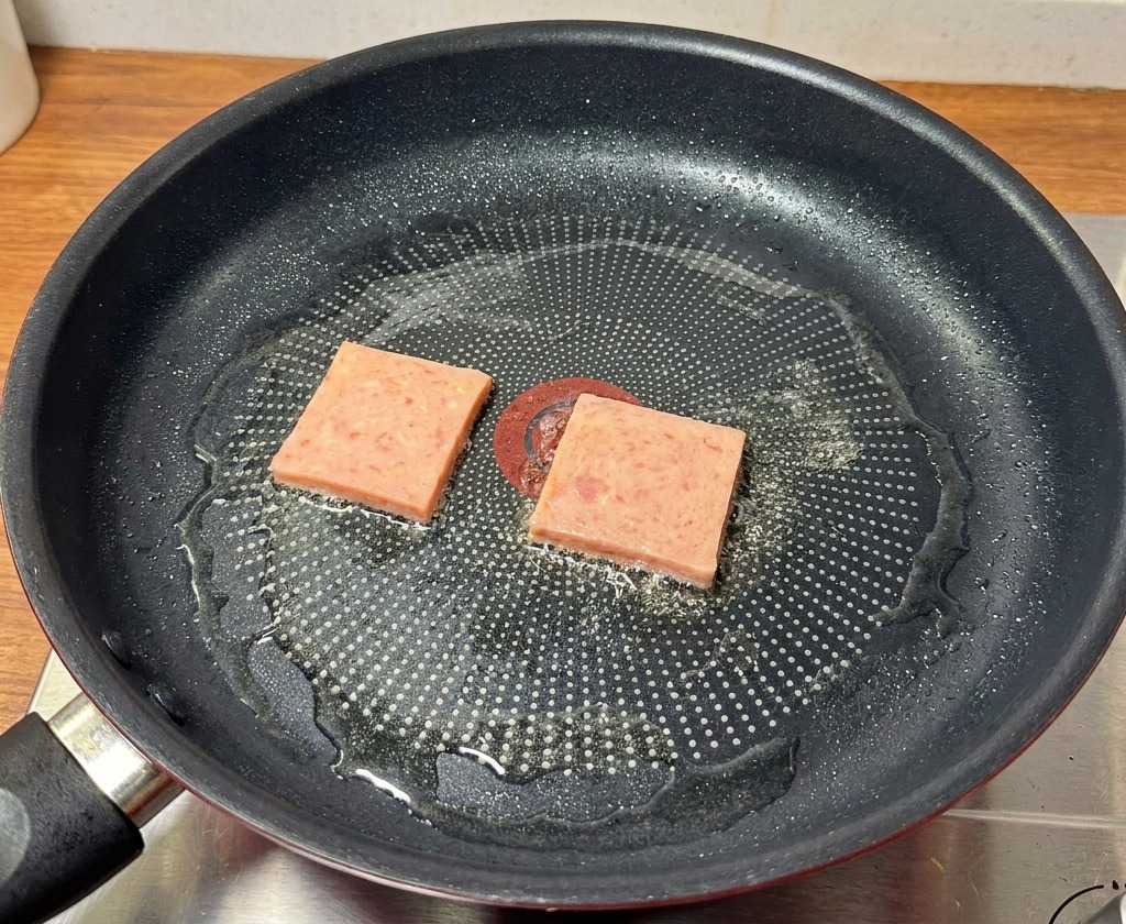 검은색 프라이팬에서 런천미트 슬라이스가 노릇노릇하고 바삭하게 구워지고 있는 모습.