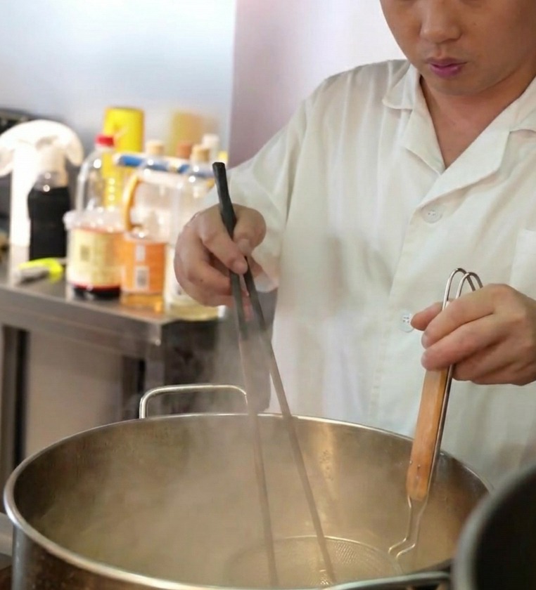 셰프가 채망에 담긴 신선한 라멘 면을 끓는 물이 담긴 커다란 냄비에 넣는 모습.