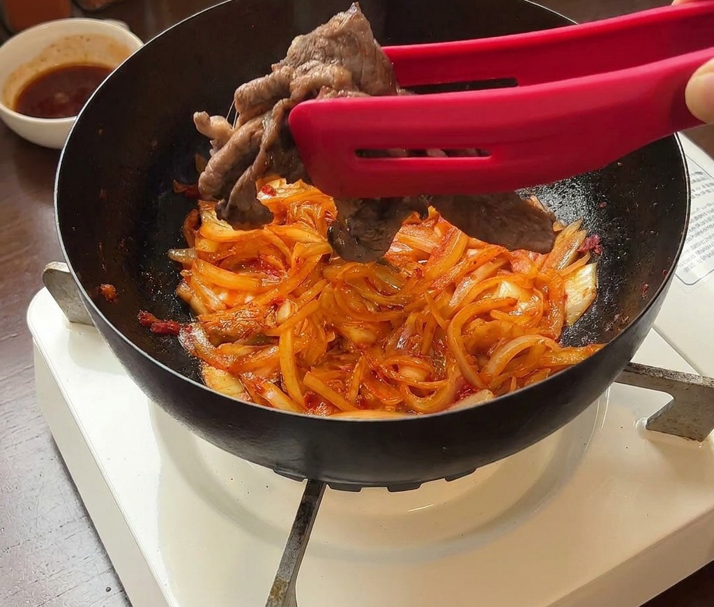 김치와 양파가 볶아진 팬에 집게를 이용해 소고기를 다시 넣는 모습.