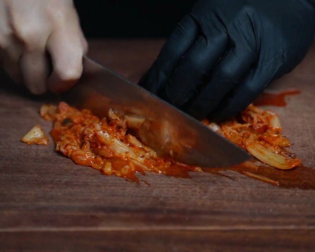 어두운 나무 도마 위에서 날카로운 칼로 붉게 잘 익은 김치를 잘게 써는 모습