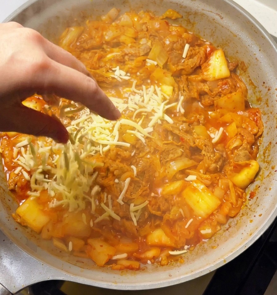 보글보글 끓는 김치볶음 재료 위에 하얀 모짜렐라 치즈를 듬뿍 뿌리는 모습.