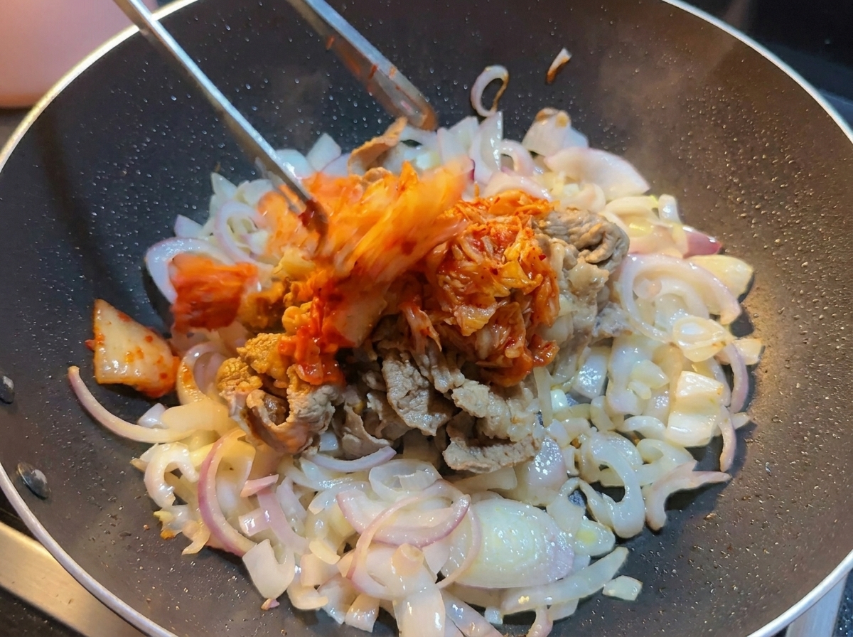우삼겹과 양파가 담긴 웍에 집게로 먹기 좋게 다진 배추김치를 넣는 장면.
