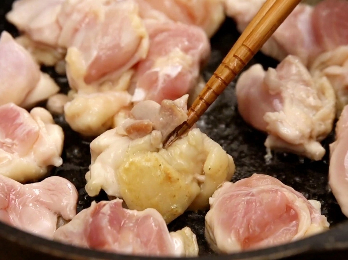 어두운 색 프라이팬에서 한입 크기의 닭고기를 젓가락으로 뒤집으며 노릇하게 볶는 모습.