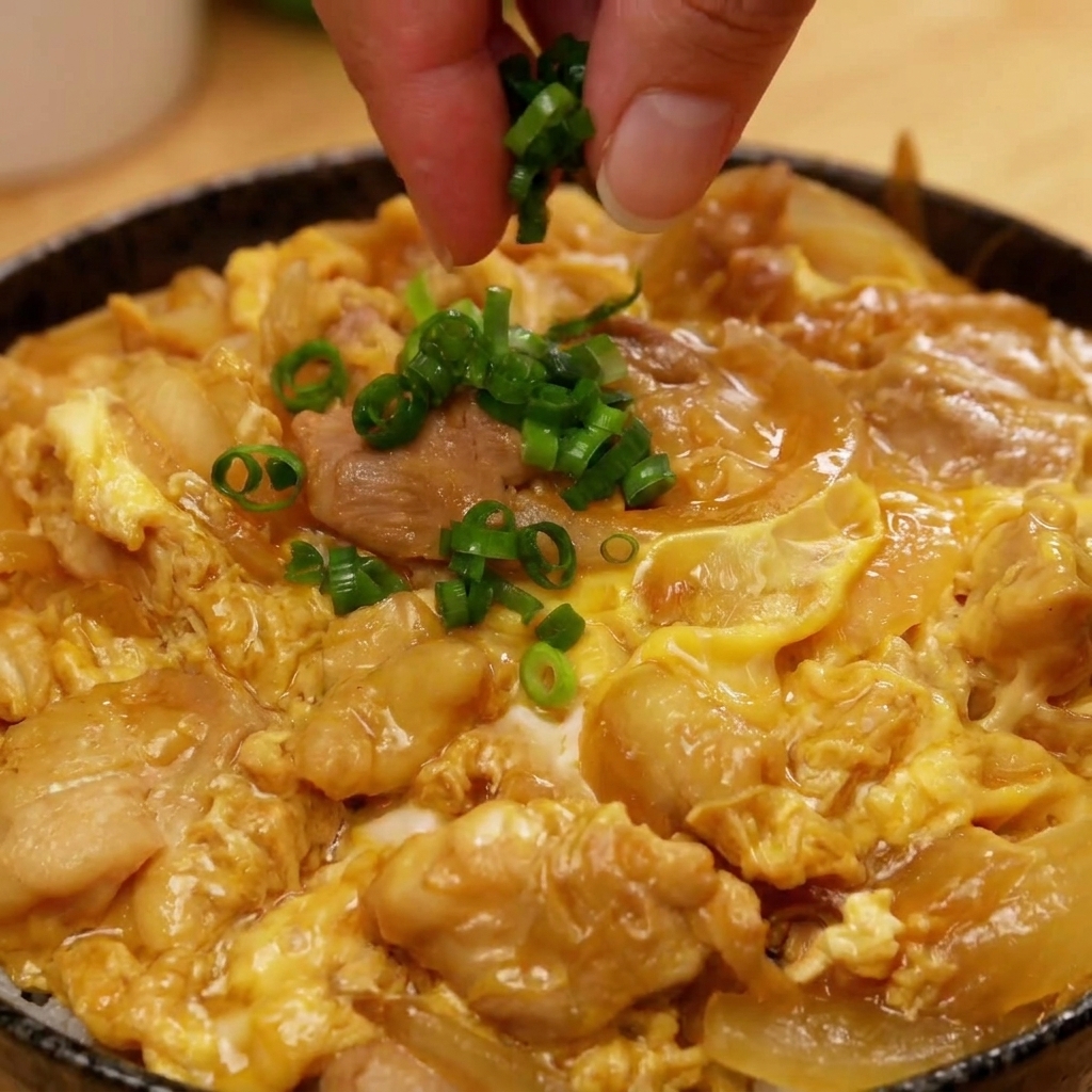 완성된 오야코동 중앙에 잘게 썬 쪽파를 뿌려 마무리하는 손의 모습.