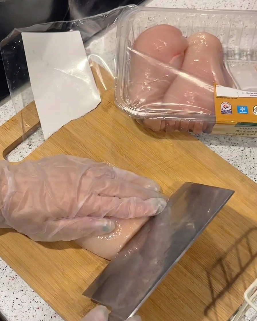 장갑을 낀 손으로 대나무 도마 위에서 닭가슴살을 얇게 포 뜨듯 써는 모습