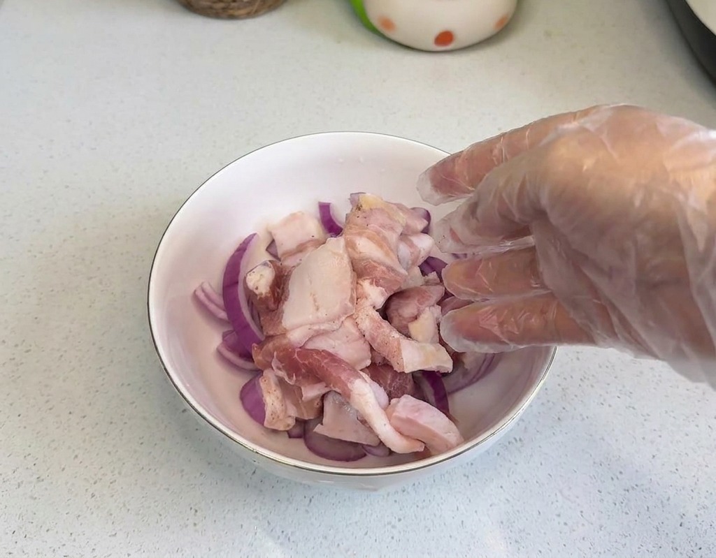 투명한 위생장갑을 낀 손으로 적양파 링이 담긴 볼에 생삼겹살을 올리는 모습.