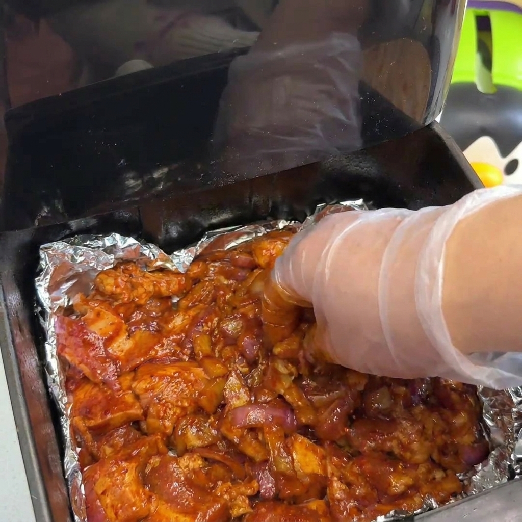 위생장갑을 낀 손으로 에어프라이어 바스켓 안에 양념된 고기와 양파를 겹치지 않게 넓게 펴주는 모습.