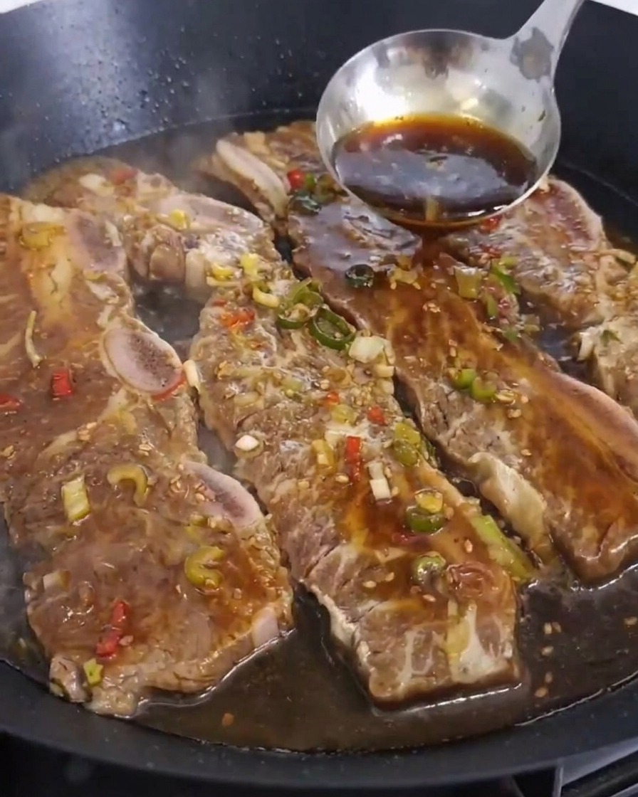 팬에서 익어가는 갈비 위로 국자를 이용해 진한 갈색 양념장을 넉넉히 붓는 장면