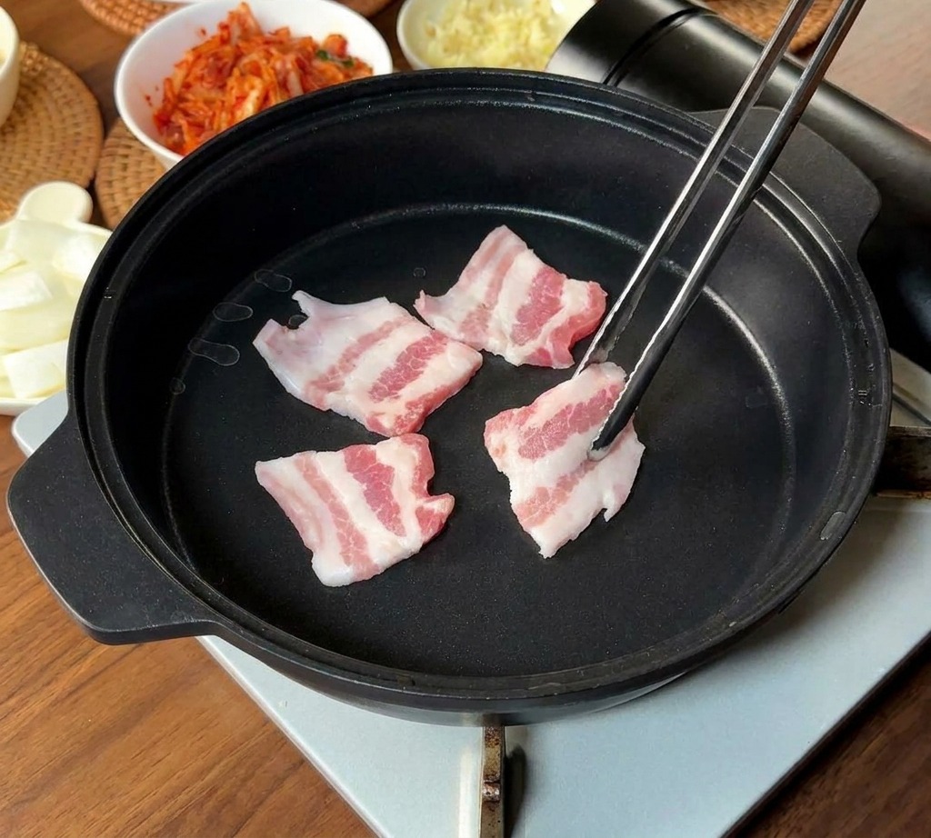 긴 금속 젓가락으로 마블링이 좋은 얇은 삼겹살을 검은 팬에 올리는 모습