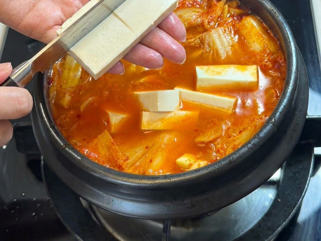 끓고 있는 매콤한 찌개 위로 도마에서 썬 두부를 조심스럽게 넣는 모습.