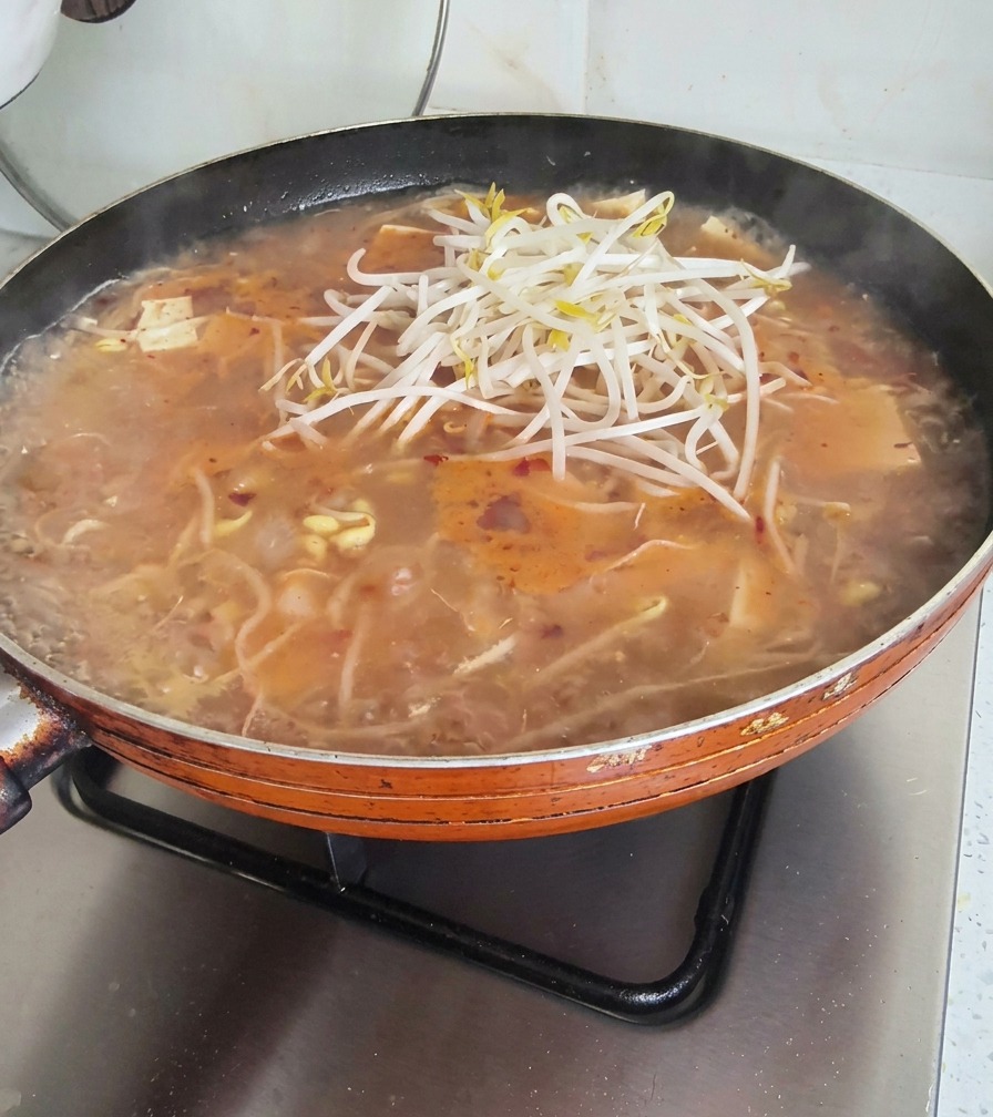끓는 찌개 냄비 위에 신선한 청경채와 남은 콩나물을 듬뿍 올리는 모습