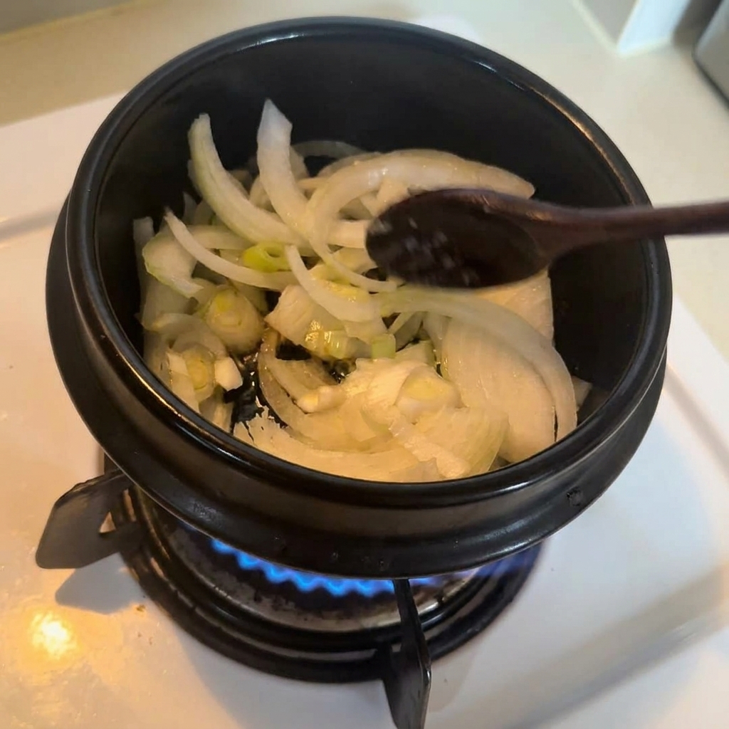 검은색 냄비 안에서 양파와 대파가 투명하게 익을 때까지 나무 주걱으로 볶는 모습.