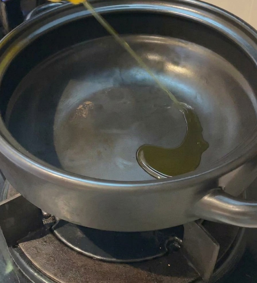 가스레인지 위 검은 뚝배기 바닥에 노란 식용유를 조금 붓고 있는 모습.