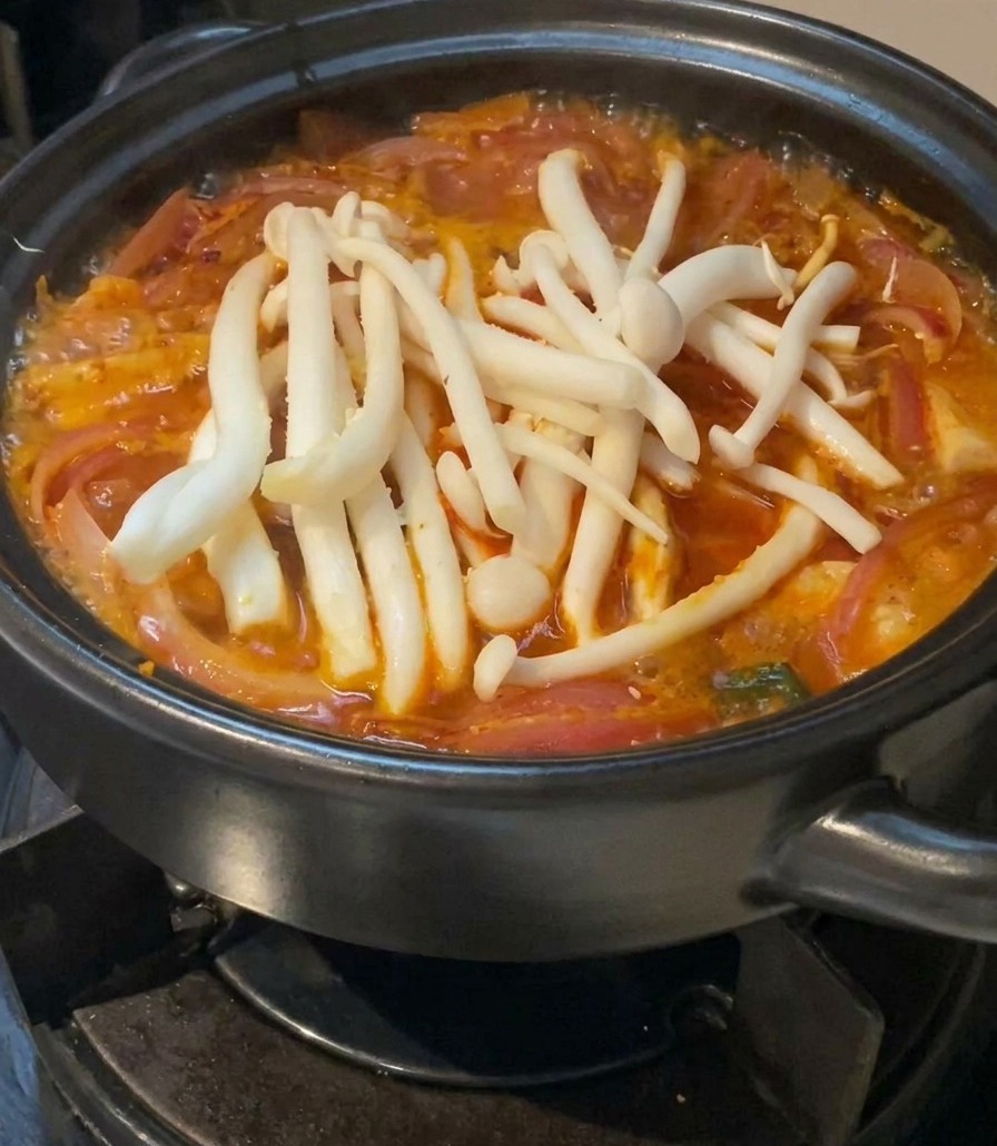 붉은 국물이 끓는 뚝배기 위에 하얀 만가닥버섯 한 뭉치를 올린 모습.